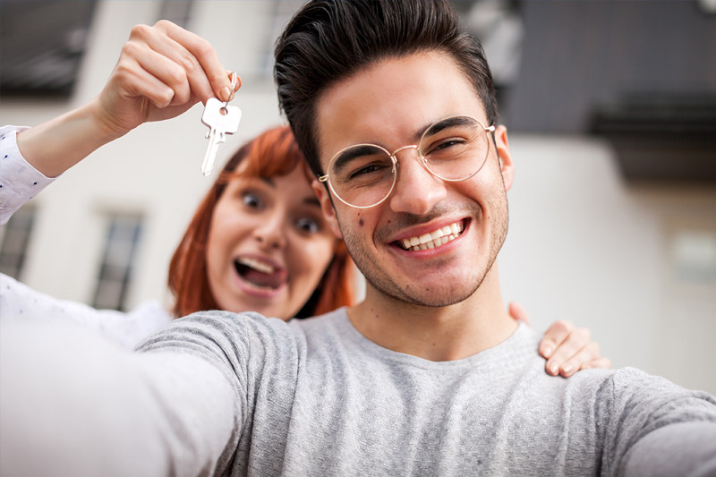 A smiling young man takes a selfie while a woman behind him excitedly holds up keys, suggesting they have just acquired a new home. Both appear happy and celebratory.